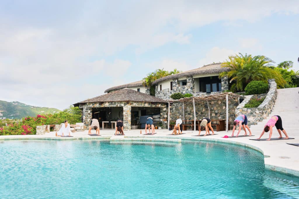 Guests participating in a yoga class beside a large pool with a stone villa in the background.