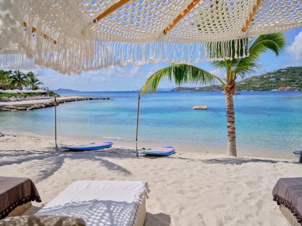View from a shaded beach lounger looking out to turquoise water with paddleboards and a palm tree.