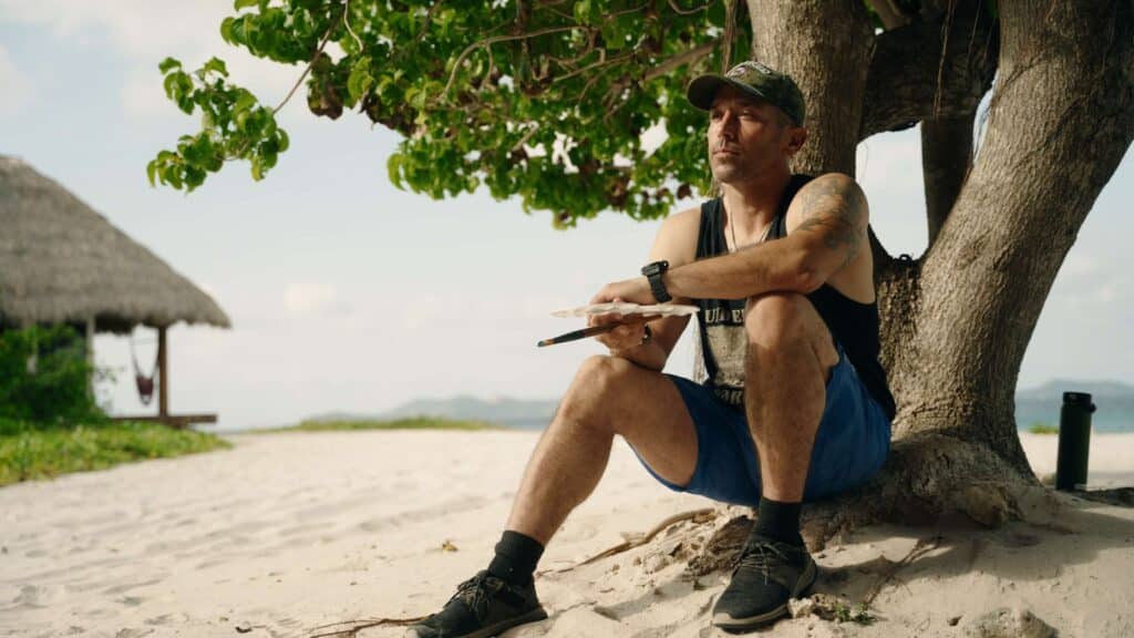 A participant sits beneath a tree on the beach holding a paintbrush and palette, reflecting during a guided creative exercise.