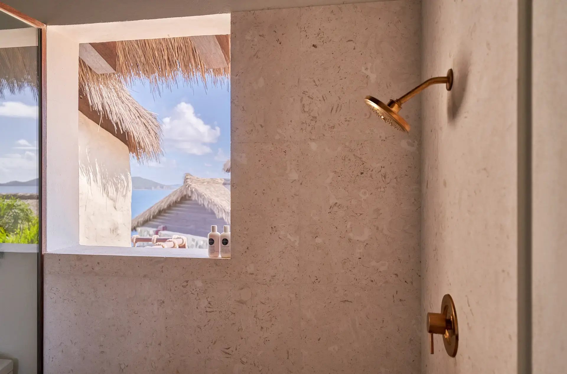 Shower with gold fixtures and window framing thatched-roof view.