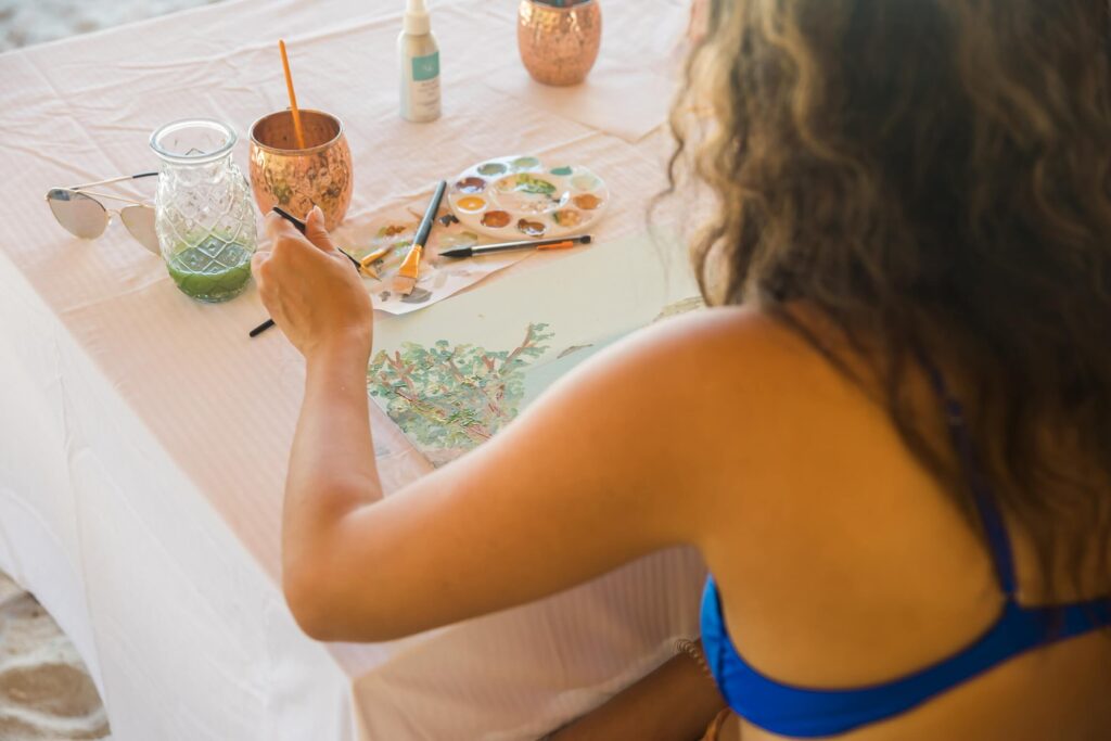 Guest painting at a table with brushes and a palette during a beachside art session.