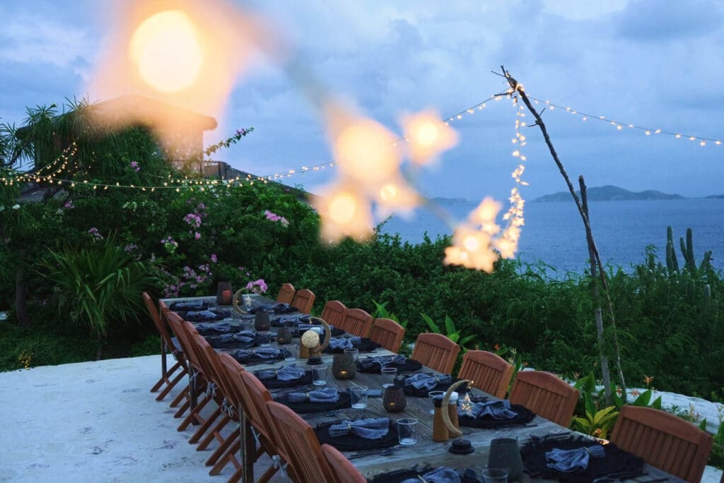 Long outdoor dining table set with string lights at dusk overlooking the ocean.