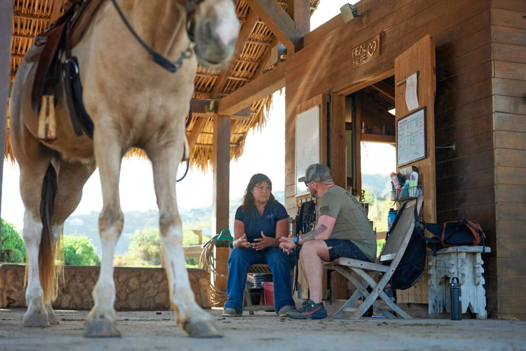 A veteran and facilitator sit in conversation outside a rustic barn while a rescued horse stands nearby at Indy’s Redemption Ranch.