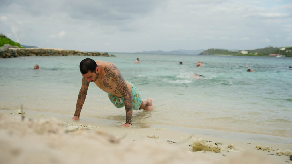 A participant performs a bodyweight exercise at the shoreline while others swim in the ocean during a group wellness session.