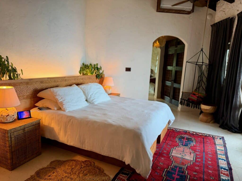 Wide view of a tranquil island bedroom at The Aerial, BVI featuring a king bed with white linens, natural woven decor, warm lighting, and a Zen Vein Healing Device positioned beside the bed for nightly wellness and recovery.