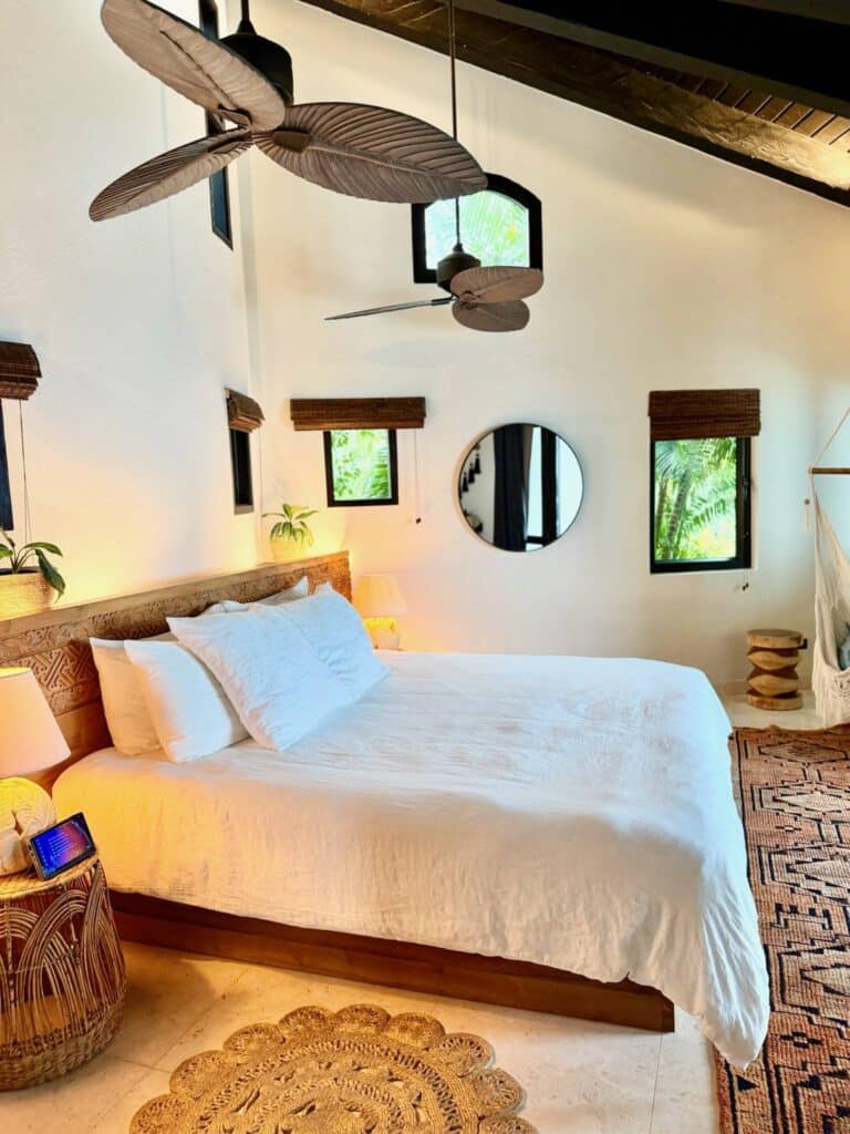 Airy island bedroom at The Aerial, BVI with high ceilings, sculptural ceiling fans, tropical greenery views, white linen bedding, and a Zen Vein Healing Device placed next to the bed as part of the resort’s holistic wellness experience.