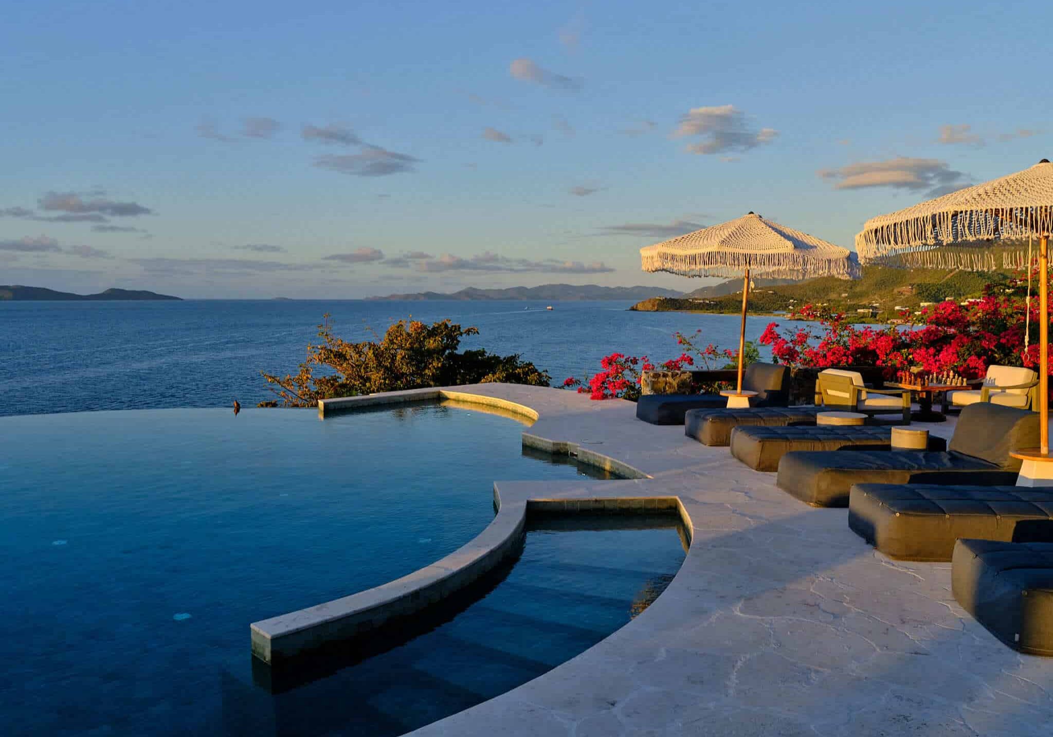 Infinity pool with lounge chairs and umbrellas facing the ocean at sunset.