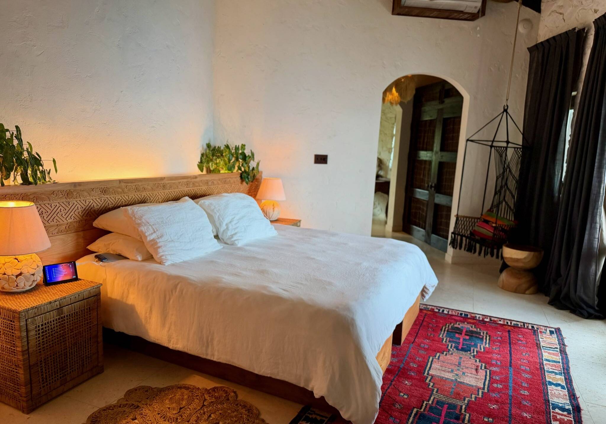 Wide view of a tranquil island bedroom at The Aerial, BVI featuring a king bed with white linens, natural woven decor, warm lighting, and a Zen Vein Healing Device positioned beside the bed for nightly wellness and recovery.